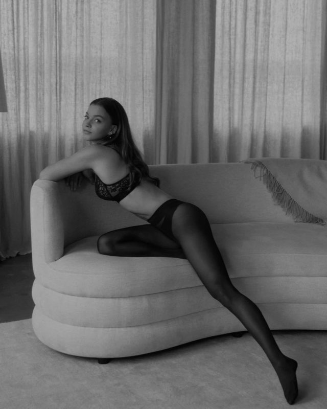 Black-and-white photograph of a model in lace lingerie and sheer tights leaning over a curved sofa